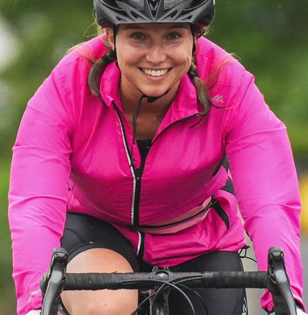 Frau sitzt mit zwei Zöpfen und Helm auf dem Fahrrad, lächelt und wird vom Wind von hinten unterstützt